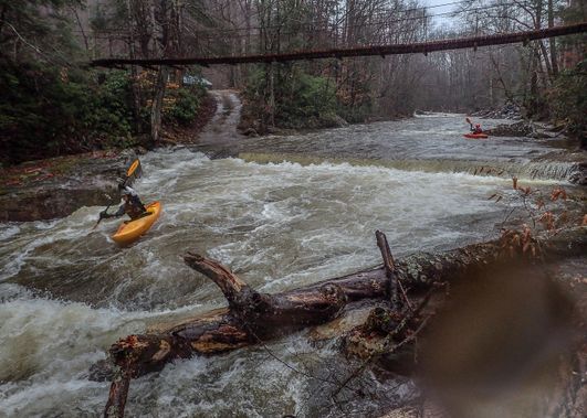 Kayakers paddle under a suspension footbridge on the North Fork of the Cherry River