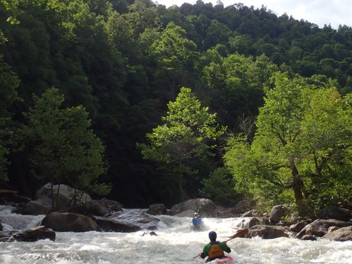 A kayaker waits in a pool while another navigates the last part of the rapid