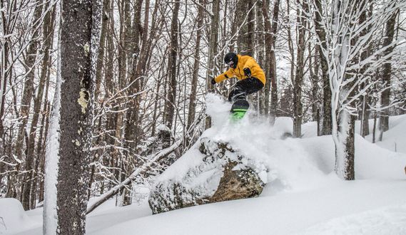 A snowboarder jumps off of a powder covered rock